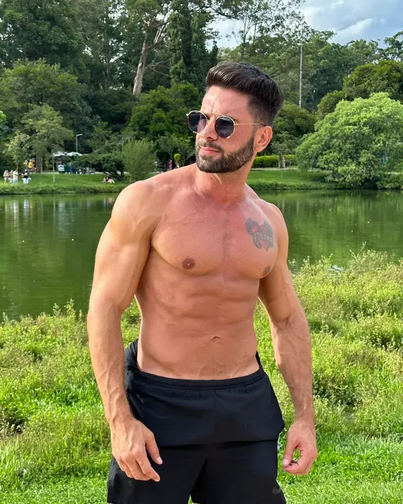 Homem sem camisa, de óculos escuros, à beira de um lago